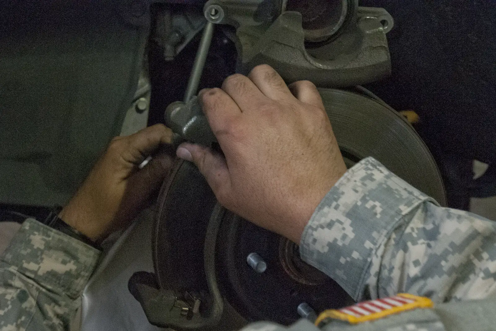 Mechanic's hands at a car's front brake with the wheel off, the brake caliper unbolted and lifted above the rotor so its round piston is visible