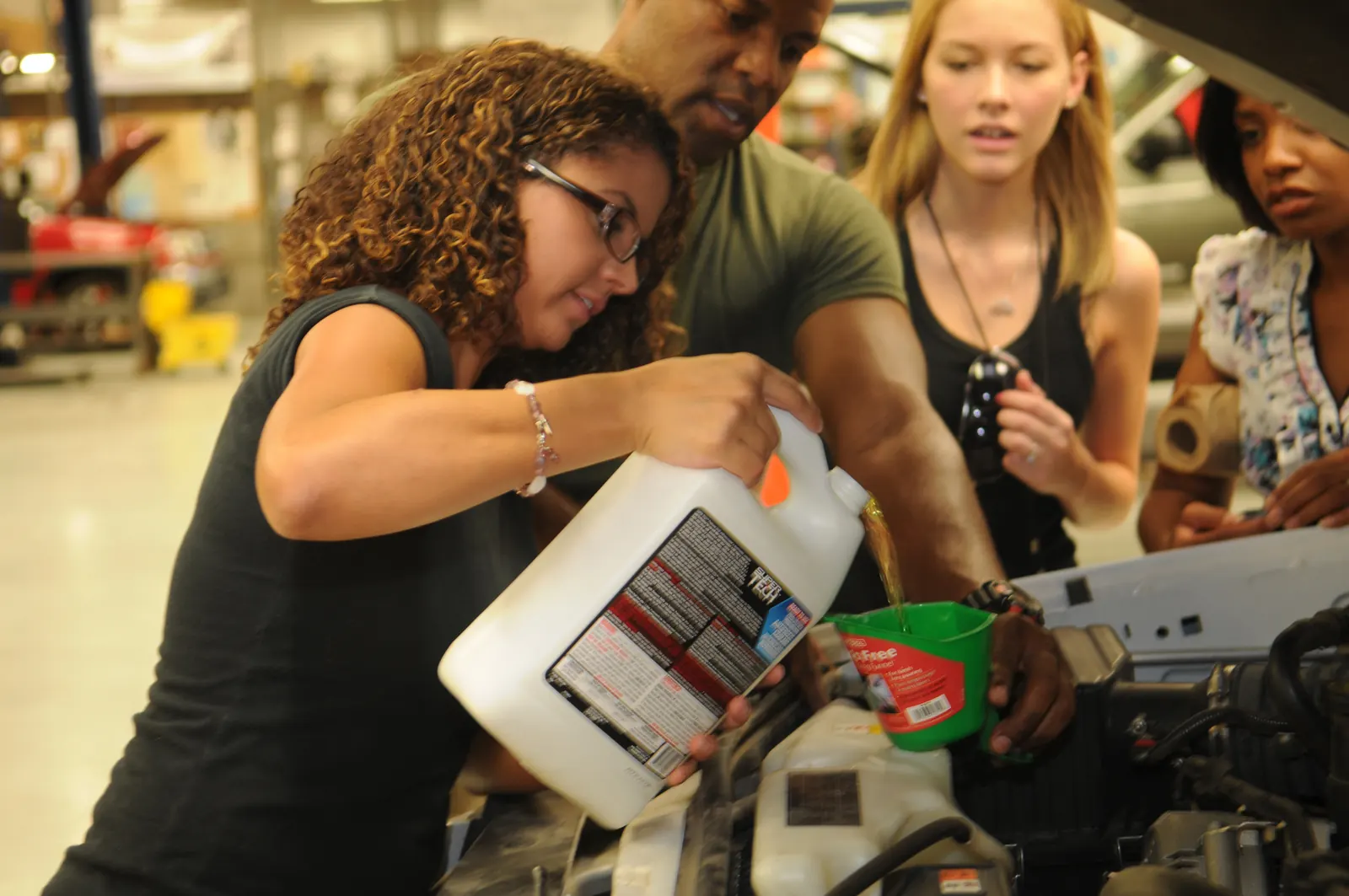 Woman pouring antifreeze from a jug through a green funnel into a car's coolant reservoir in a repair shop, with three people watching under the open hood