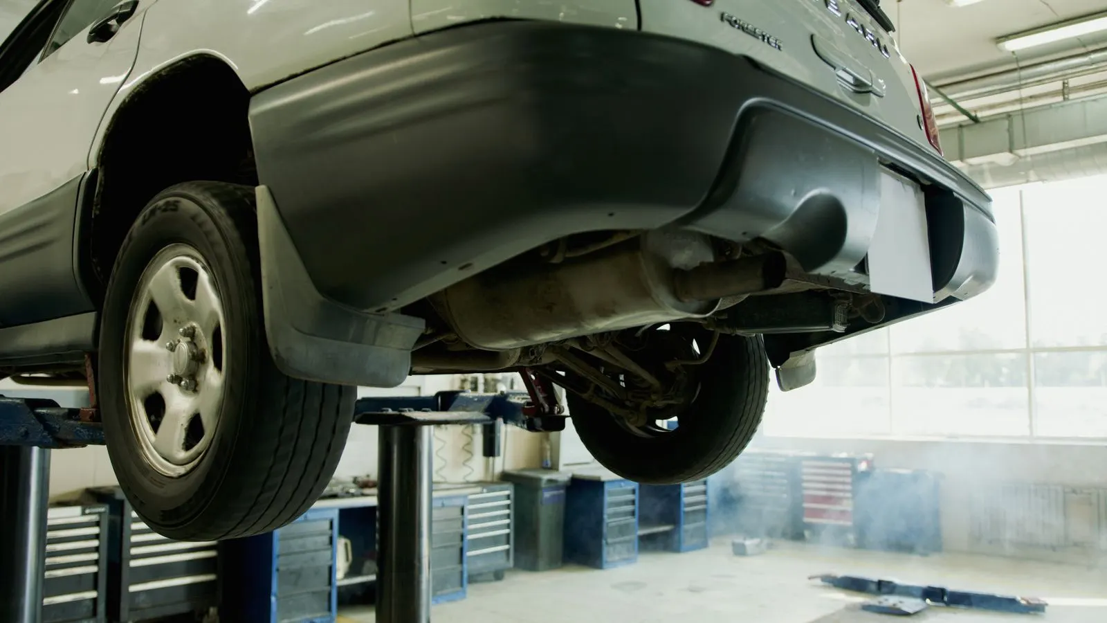 A light-colored SUV raised on a two-post lift in a repair shop, showing the underside, rear axle and exhaust where a technician inspects mounts and suspension