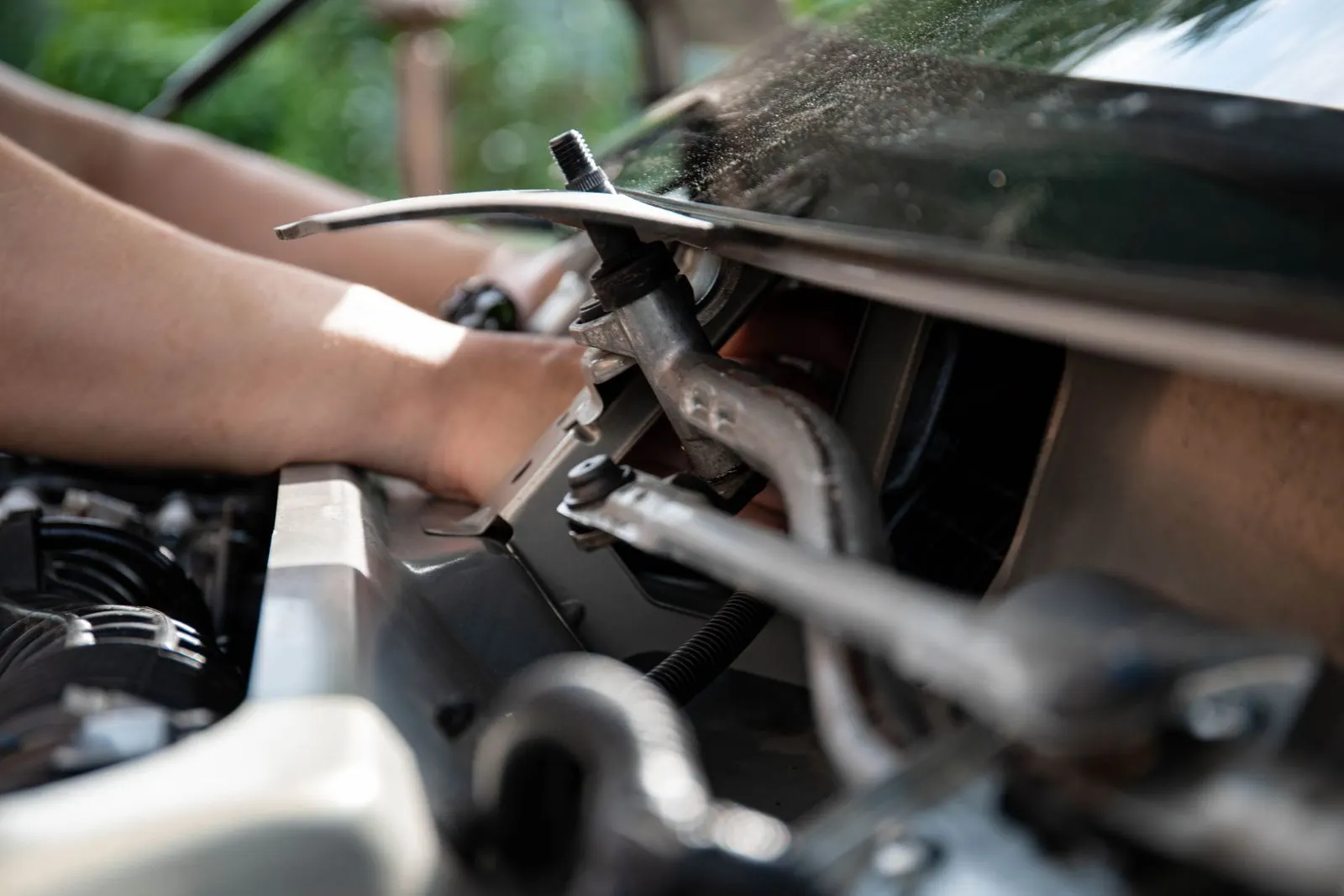 A mechanic's hands reaching under an open car hood to work on a metal bracket in the engine bay