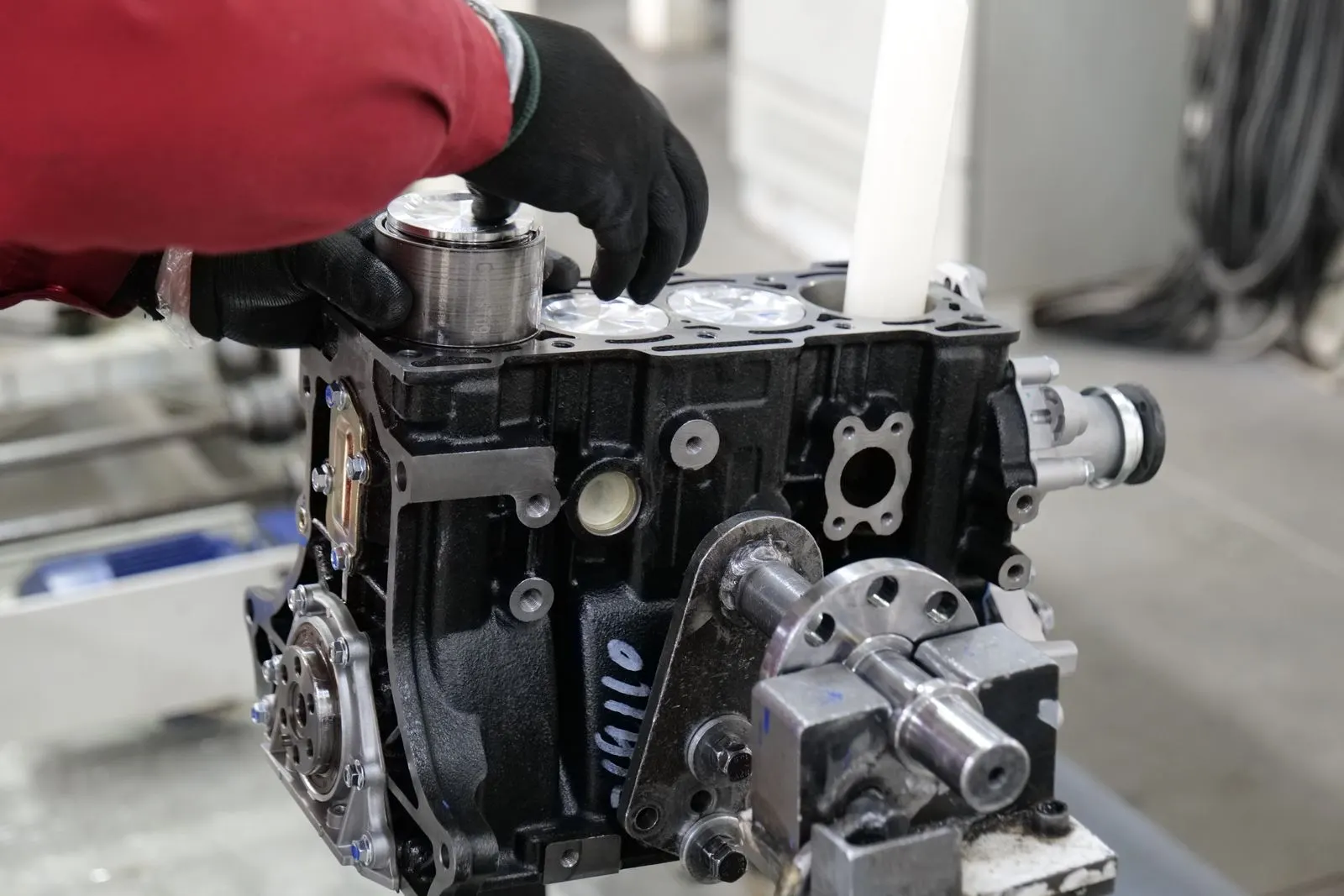 Gloved hands fitting a piston into the bore of a bare engine block mounted on an engine stand during a rebuild