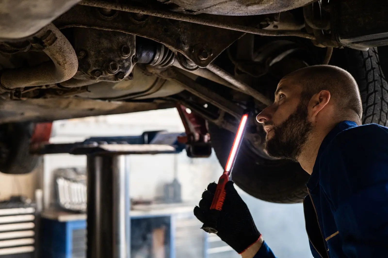 A mechanic holding a work light while inspecting the underside of a car raised on a lift