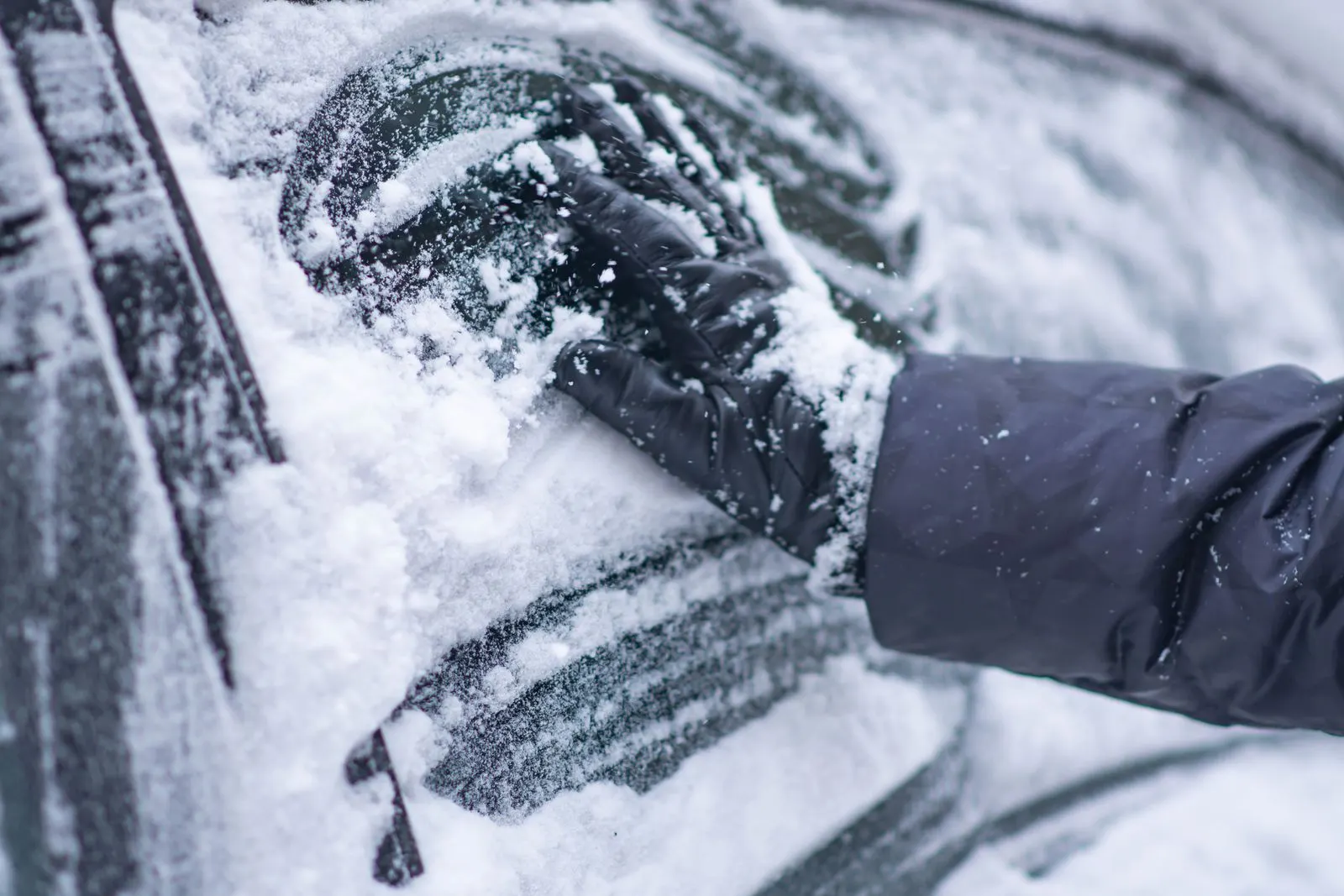 A gloved hand brushing snow off a car window on a winter day