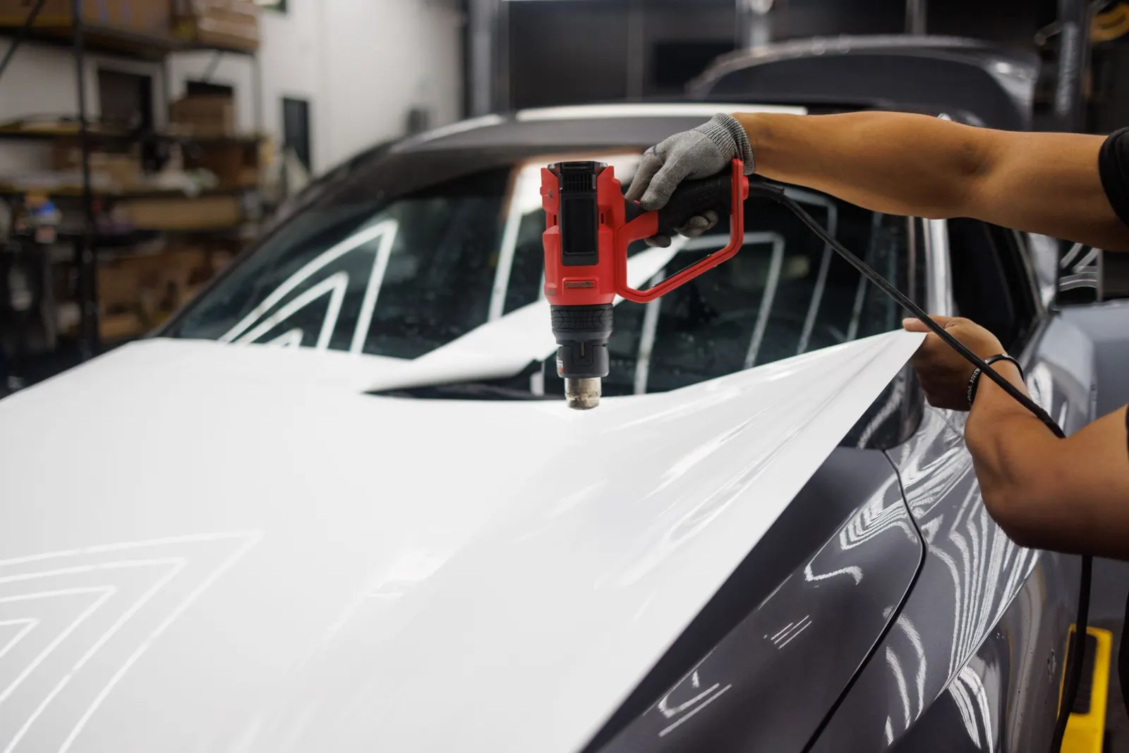 An installer's gloved hand holding a heat gun over white vinyl wrap film being stretched across the hood of a gray car in a workshop